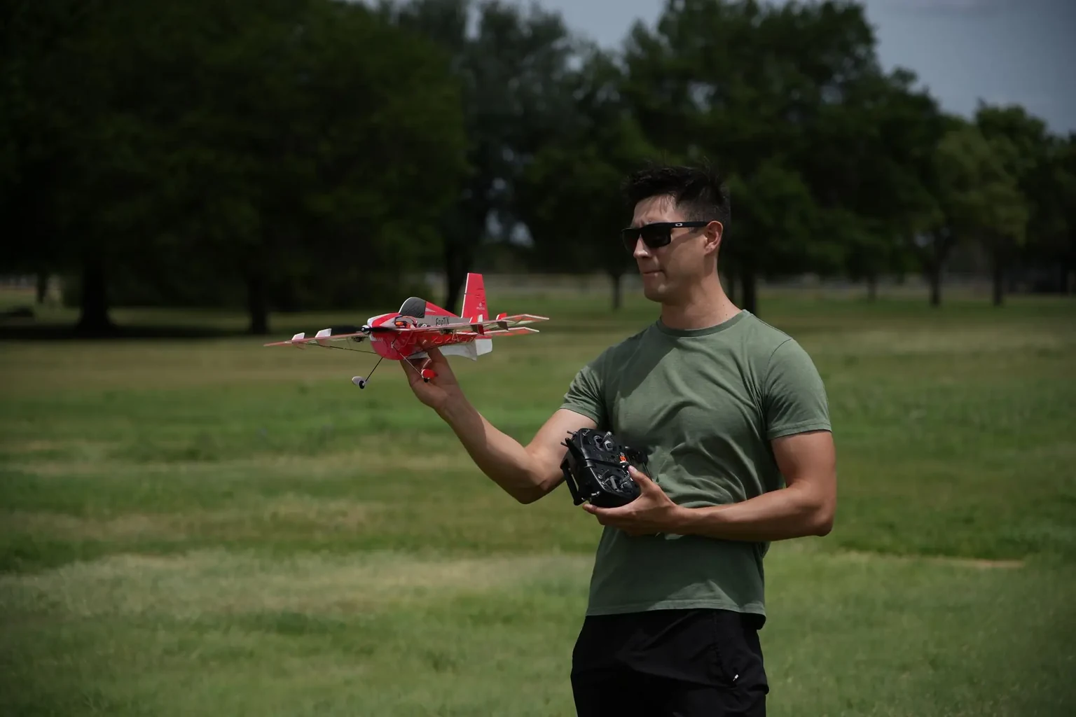 An aeromodelling pilot holding a red model aircraft and a radio controller in an open field